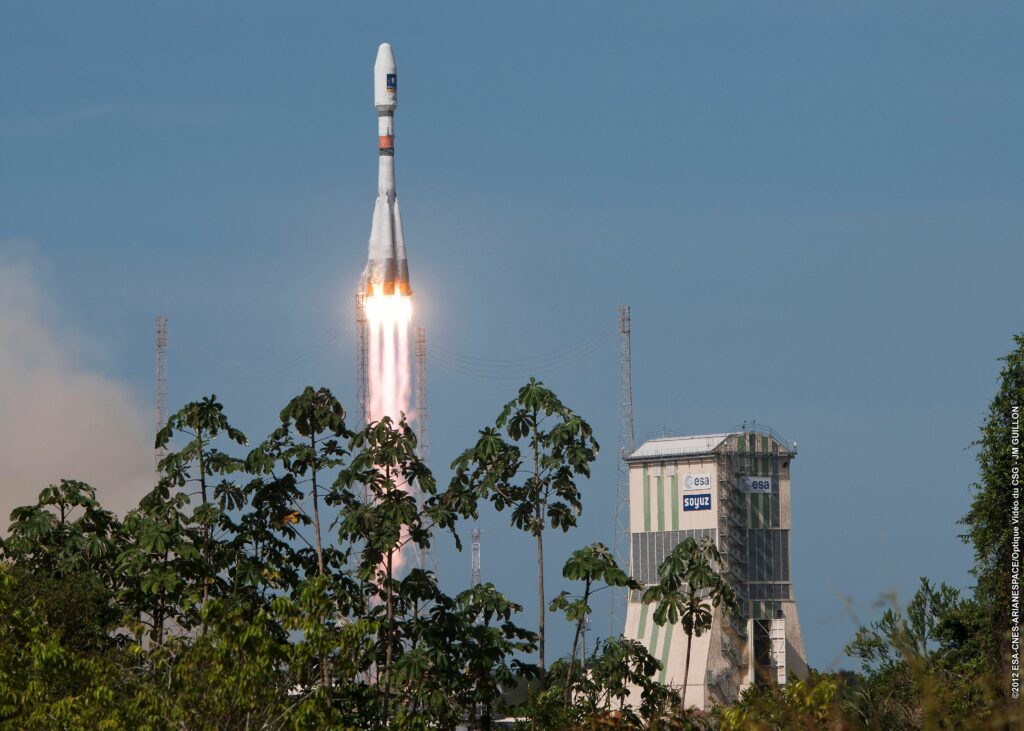 Photo d'une fusée qui s'envole du Centre Spatial Guyanais. Devant des feuilles d'arbre et au dernier plan, le tour de contrôle.